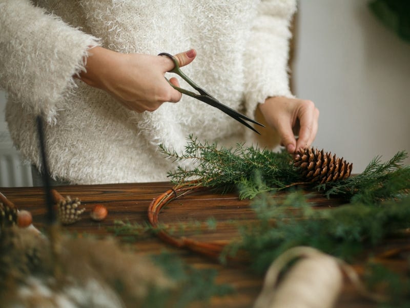 Cilvēks ar šķērēm gatavo svētku dekoru no skuju zariem un priedes čiekura uz koka galda.