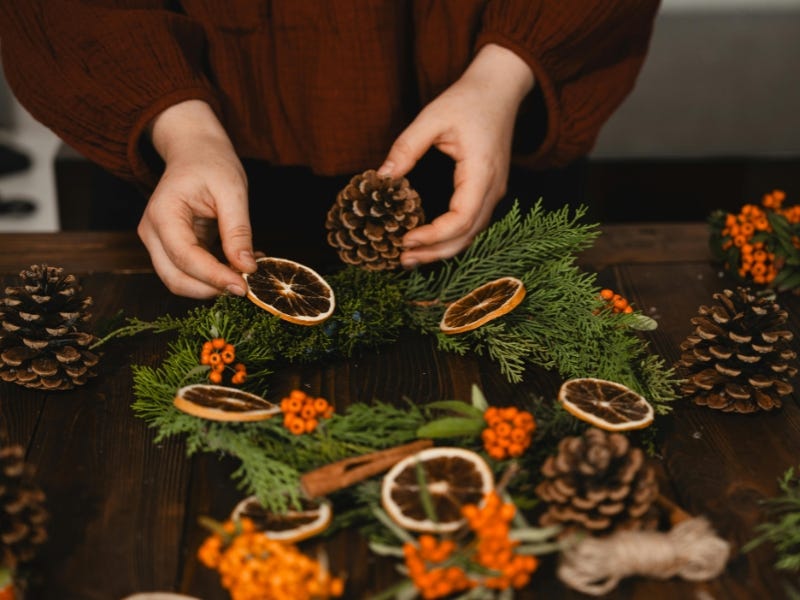 Cilvēks veido svētku vainagu no skuju zariem, kaltētiem apelsīnu šķēlītēm un čiekuriem uz koka galda.