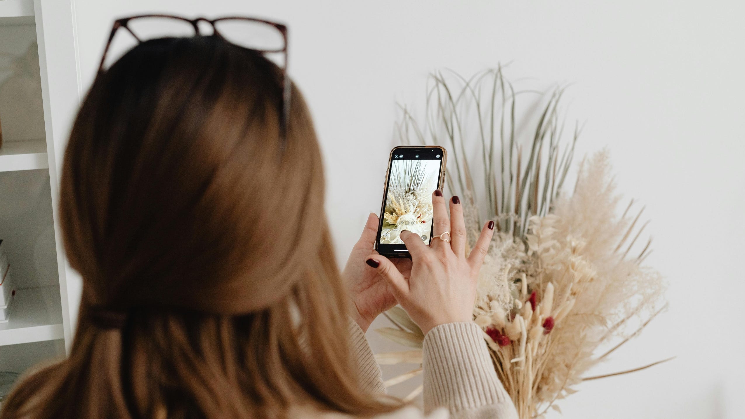 Sieviete ar telefonu fotografē dekoratīvus kaltētus augus gaišā interjerā.