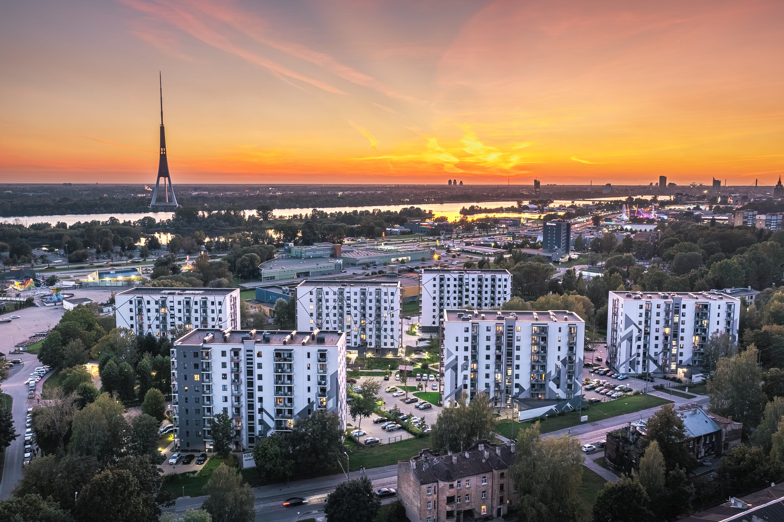 Rīgas panorāma ar daudzdzīvokļu mājām, televīzijas torni un saulrietu pār Daugavu.