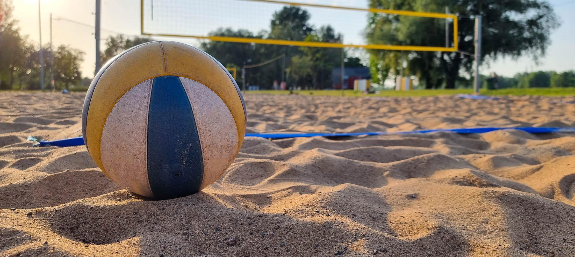 Bonava latvija Volejbola bumba smilšainā pludmales volejbola laukumā saulainā dienā. Fonā redzams tīkls, zālājs un koki, kas norāda uz āra aktivitāšu vidi