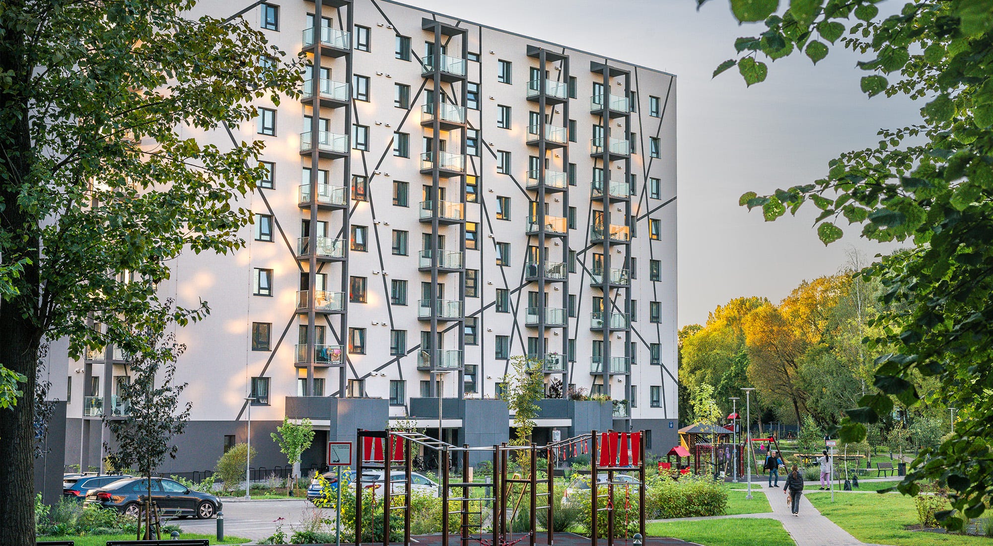 Mūsdienīgs daudzdzīvokļu nams ar rotaļu laukumu priekšplānā un zaļu, labiekārtotu vidi apkārt. Cilvēki pastaigājas un bauda mierīgu apkārtni.