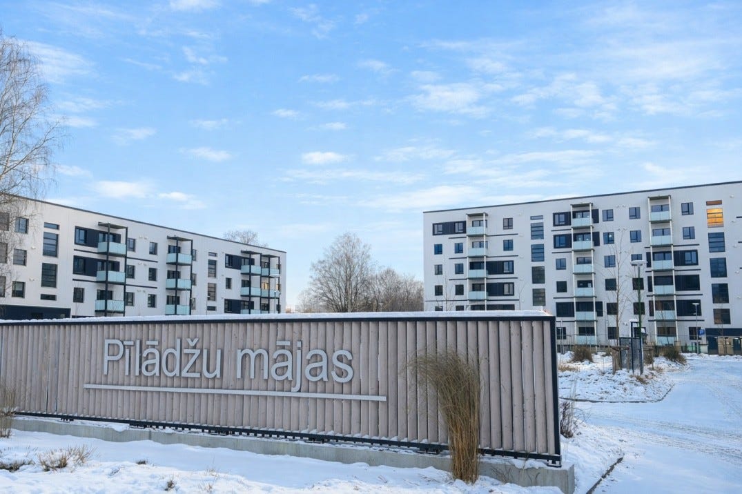 Dzīvojamais projekts “Pīlāžu mājas” ziemā – mūsdienīgas daudzdzīvokļu ēkas ar balkoniem, priekšplānā koka žogs ar projekta nosaukumu.